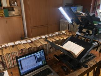 A copy stand with a camera is copying pages of a book that's sitting on a standard next to a laptop, in the background there is a large row of books to be copied.