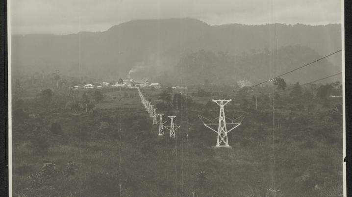 The image shows the cement factory, Indarung I, in the distance, a plume of smoke rising into the air. In the foreground, electricity lines can be seen going towards the factory. Behind the factory a mountain range creates a backdrop for the industrial scene.